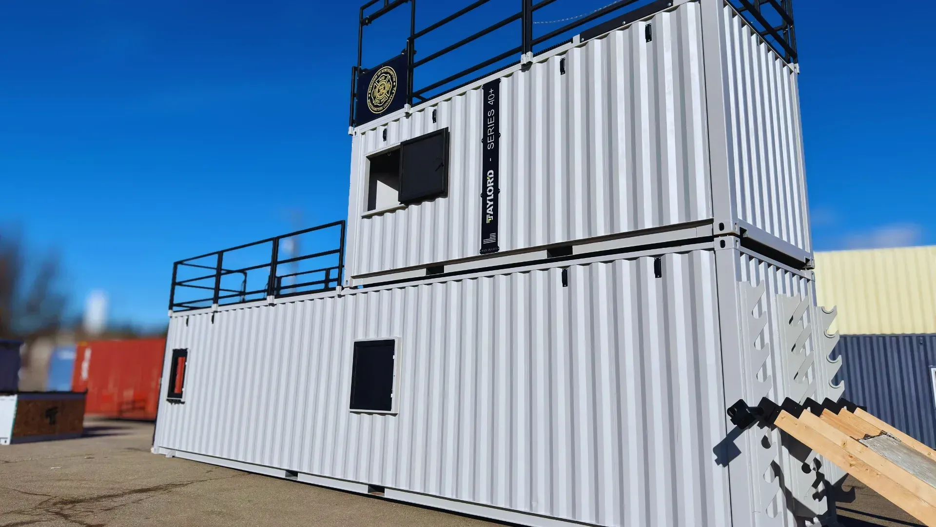 Stacked white shipping containers with black railings under a blue sky; construction site setting