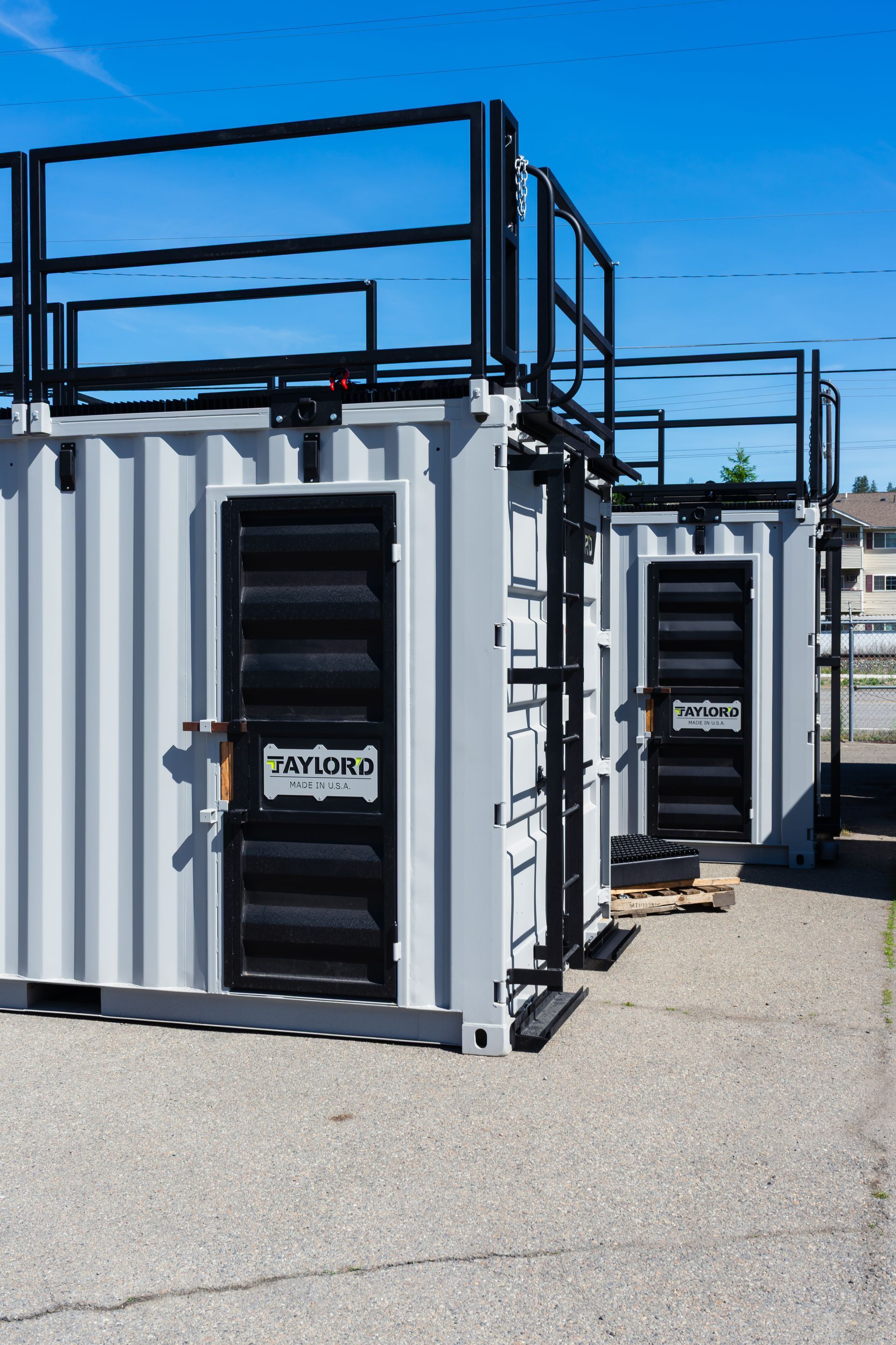 Row of white shipping containers with black metal frames on a gravel lot under a bright blue sky