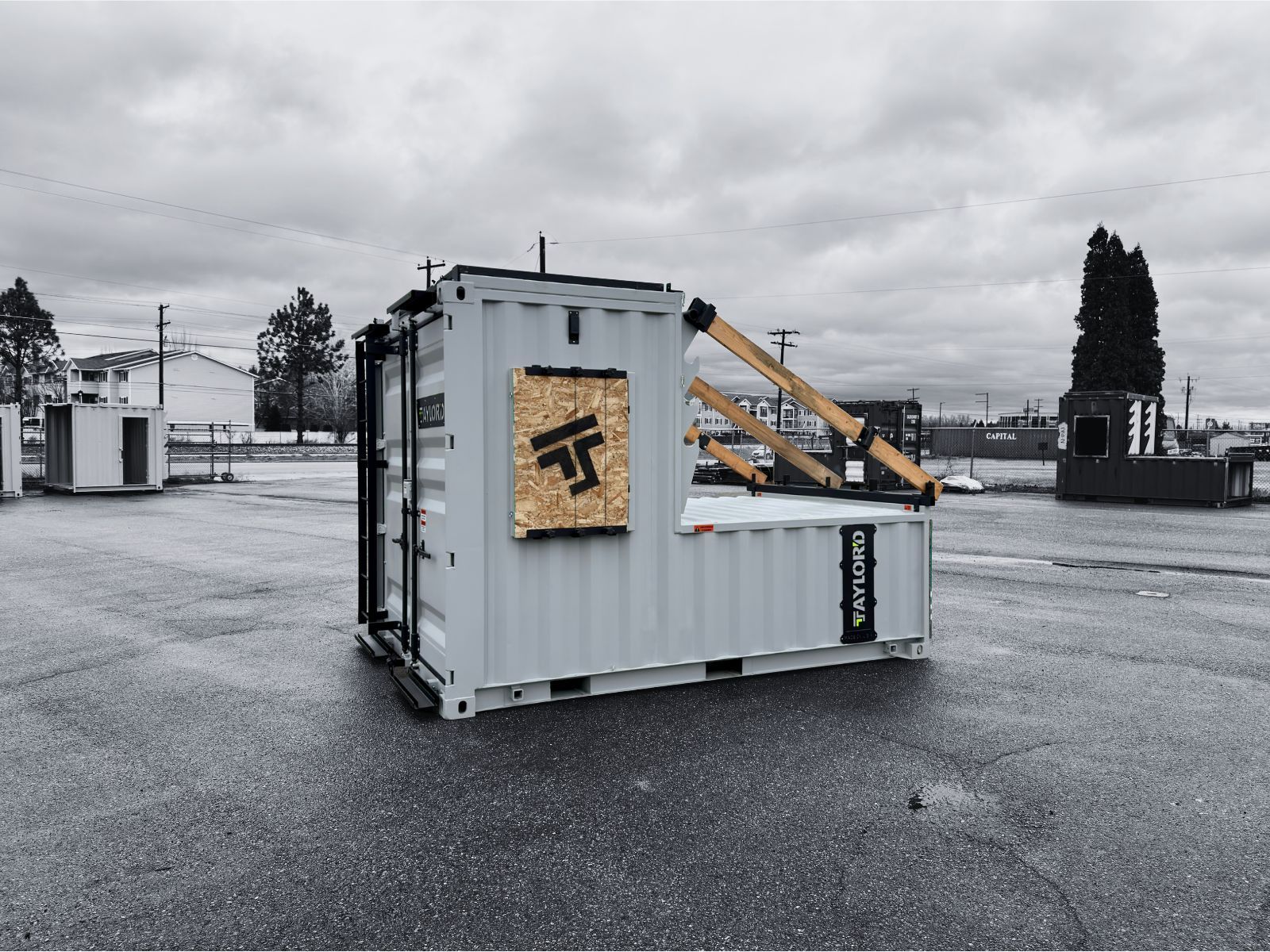 White shipping container tipped on its side in a gray industrial lot, with a wooden crate leaning against it.