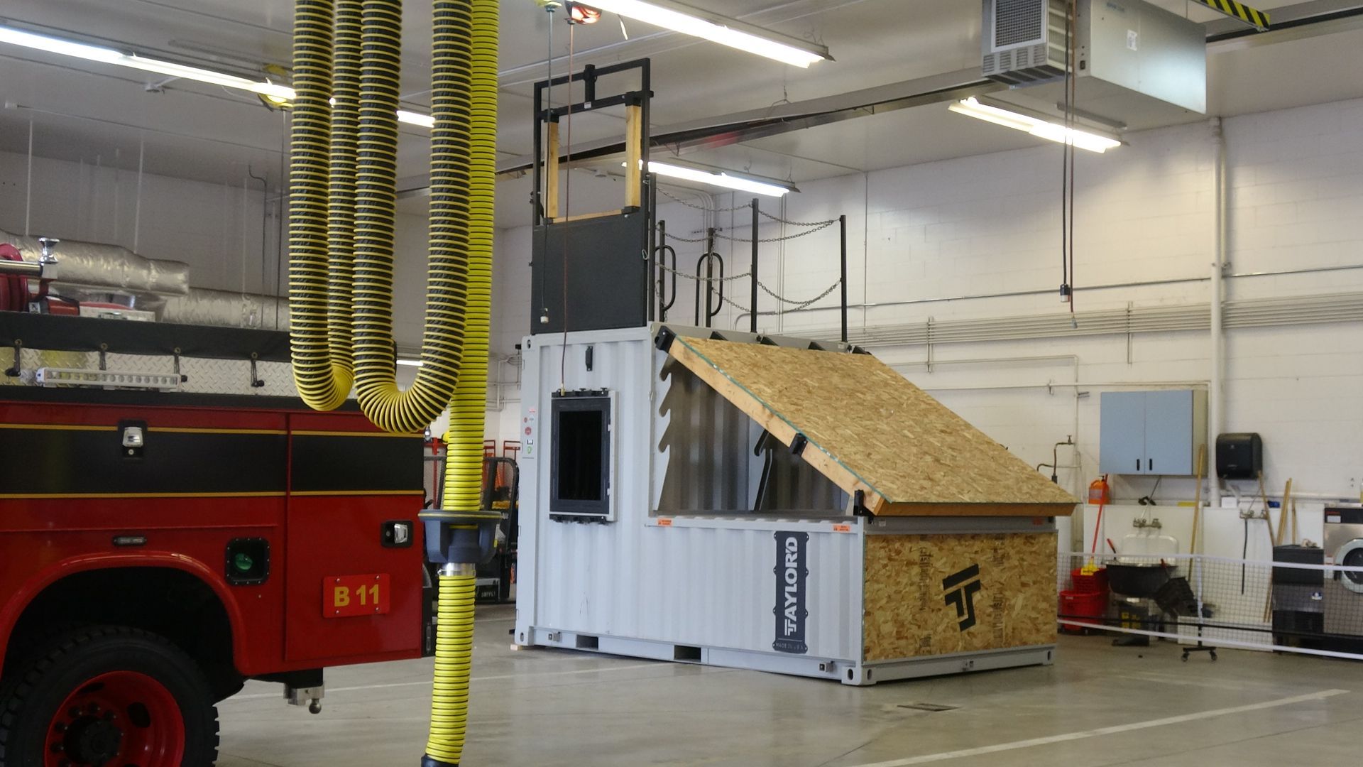 Fire truck inside a garage beside a small house mock-up with a sloped roof and yellow ventilation ducts