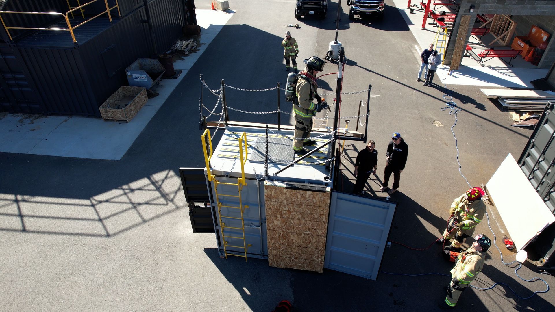 Workers moving a large blue-and-white container across a paved lot using a crane