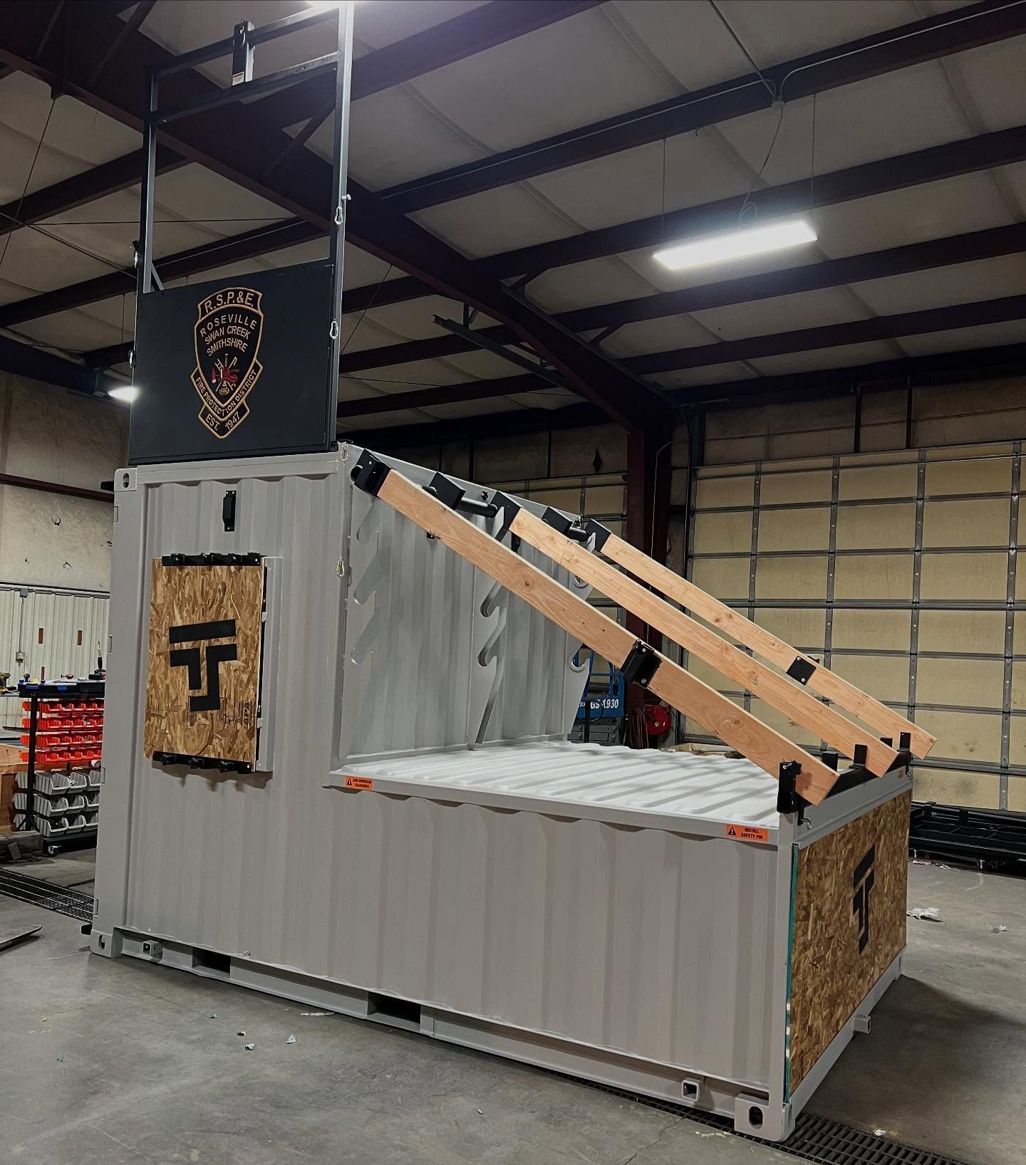 Gray shipping container in a warehouse, with a tan canopy frame and a TJ logo panel.