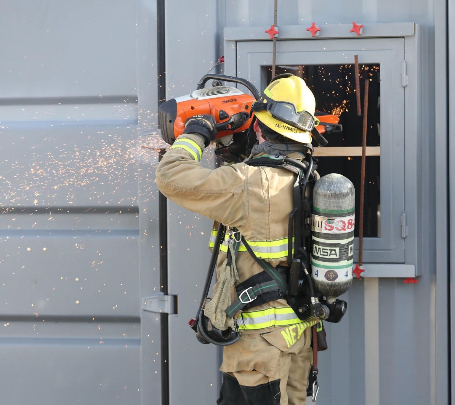 Firefighter using a chainsaw to cut through a door, with sparks flying in a gray industrial setting.