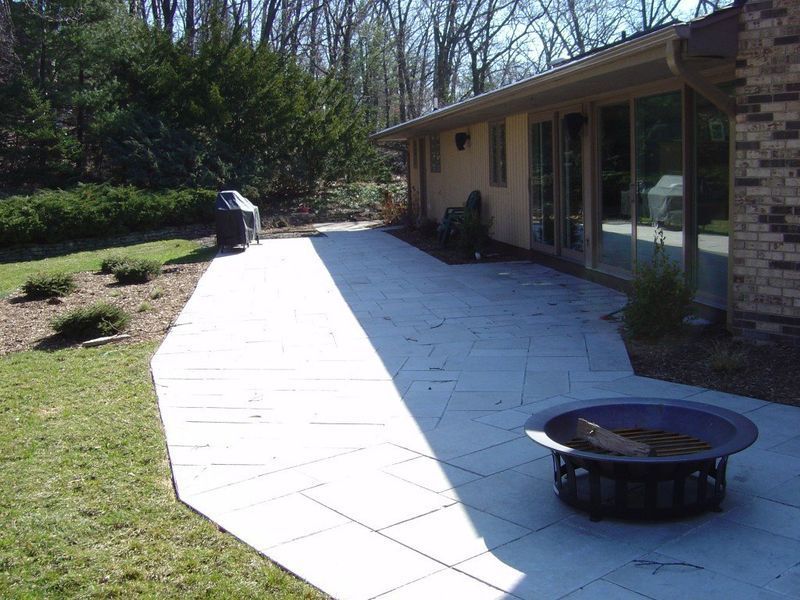 Stone patio with fire pit, grill, and house. Lawn and trees in background.