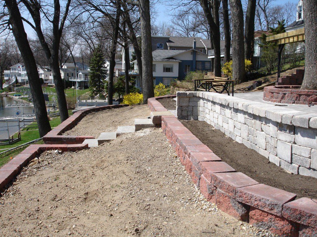 Terraced yard with retaining walls, steps, and exposed soil, overlooking a lake and houses.