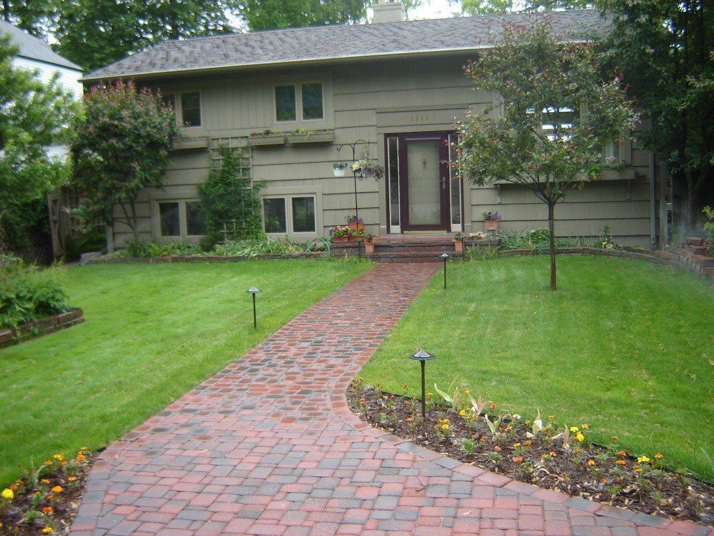 Tan two-story house with brick pathway leading to the front door, surrounded by green grass and landscaping.