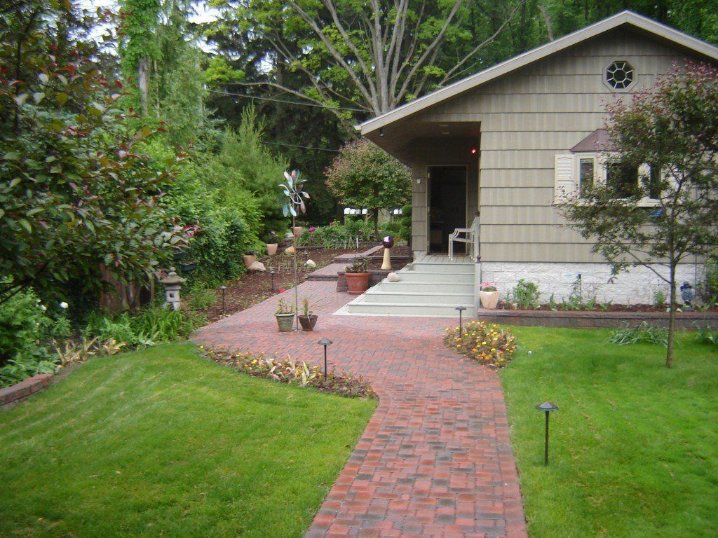 Brick pathway leads to a beige house with a covered porch, surrounded by green lawn and landscaping.