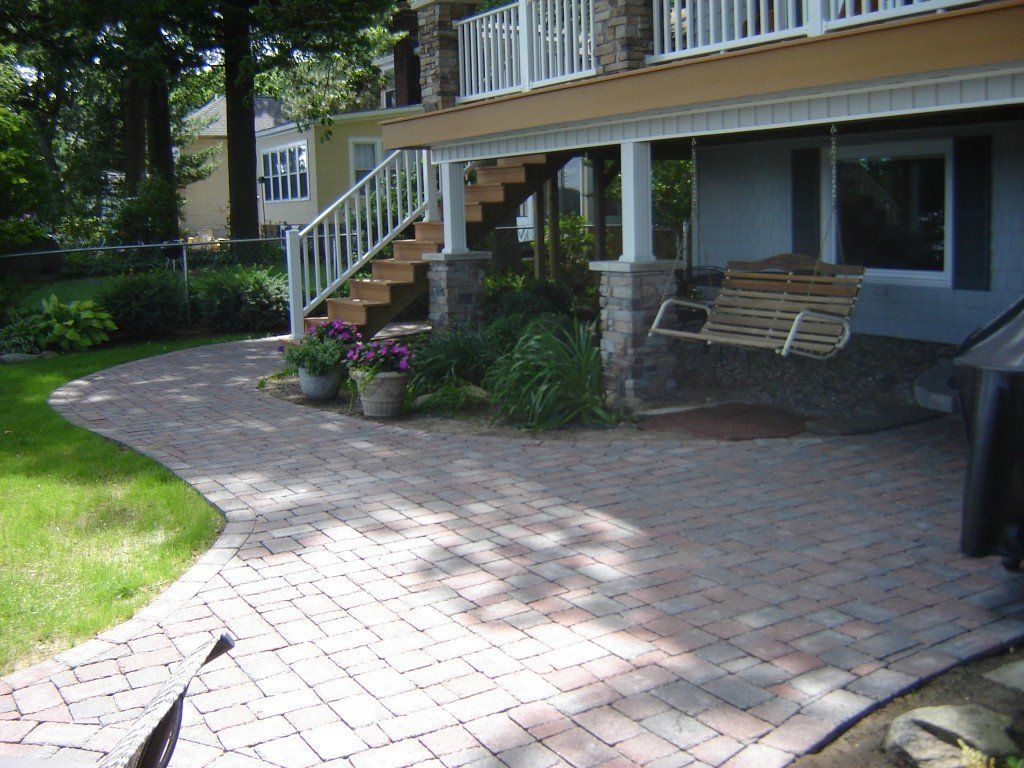 Brick patio with a swing, under a deck with stairs, next to a grassy area.