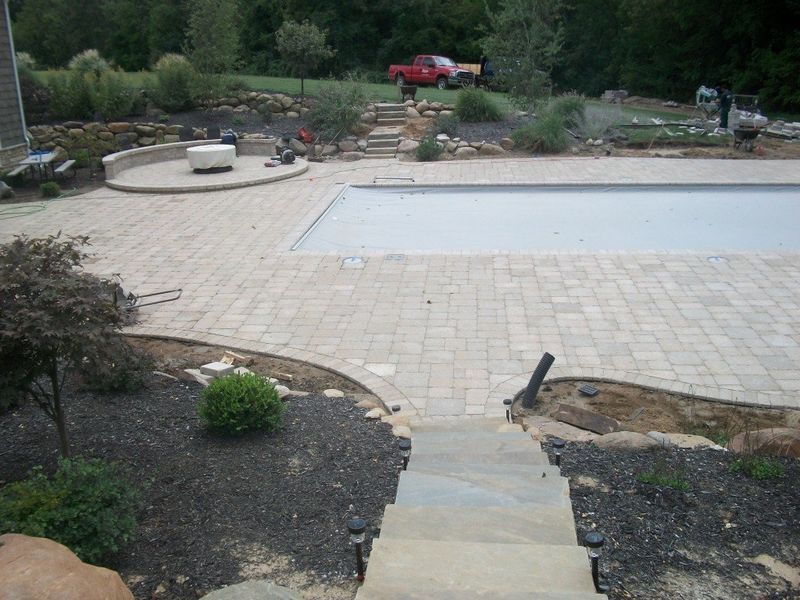 Paver patio with pool, fire pit, and stone steps leading up to red truck in the distance.
