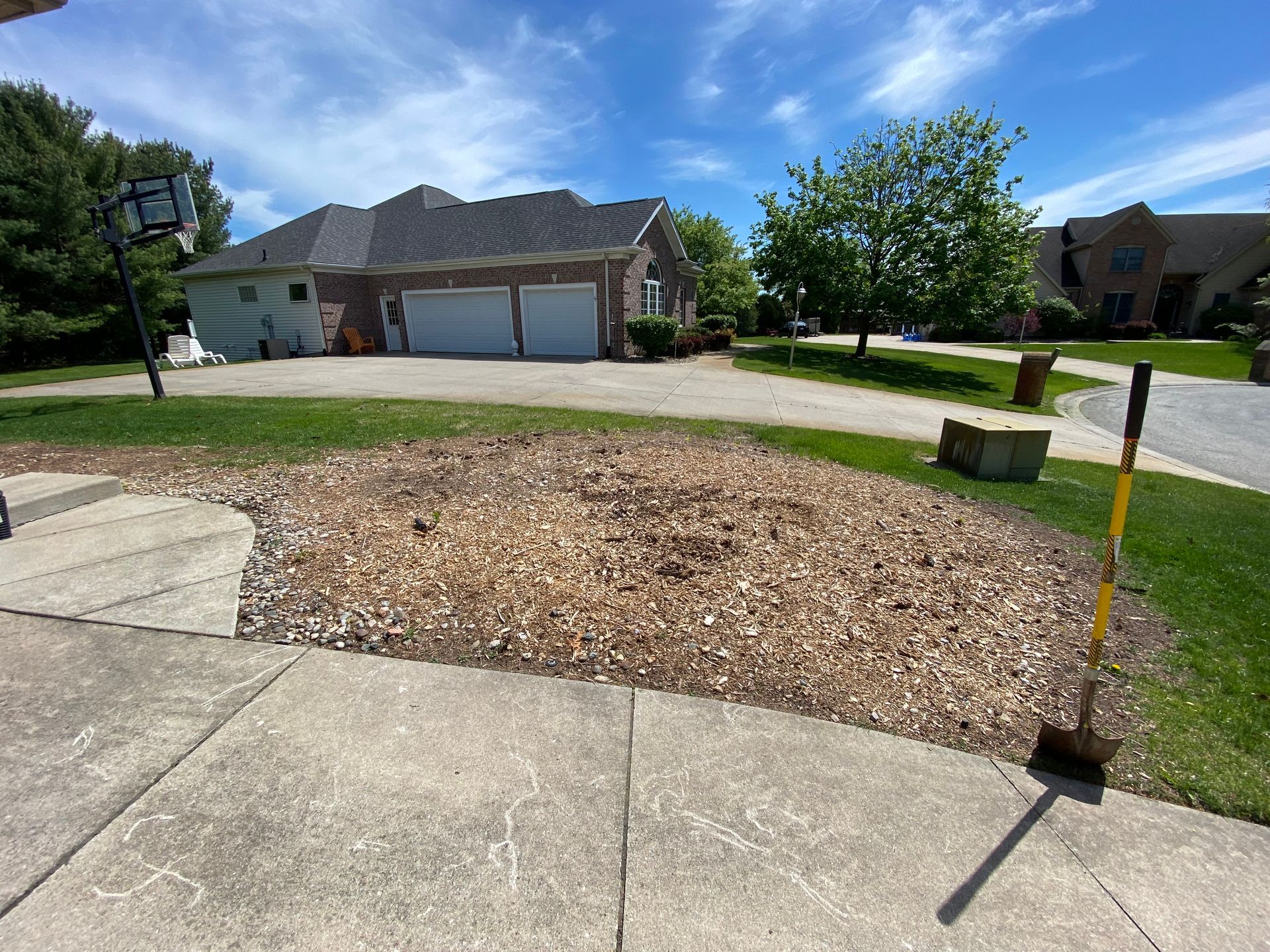 A shovel stands upright in a patch of wood chips in a residential front yard with a concrete driveway and house nearby.
