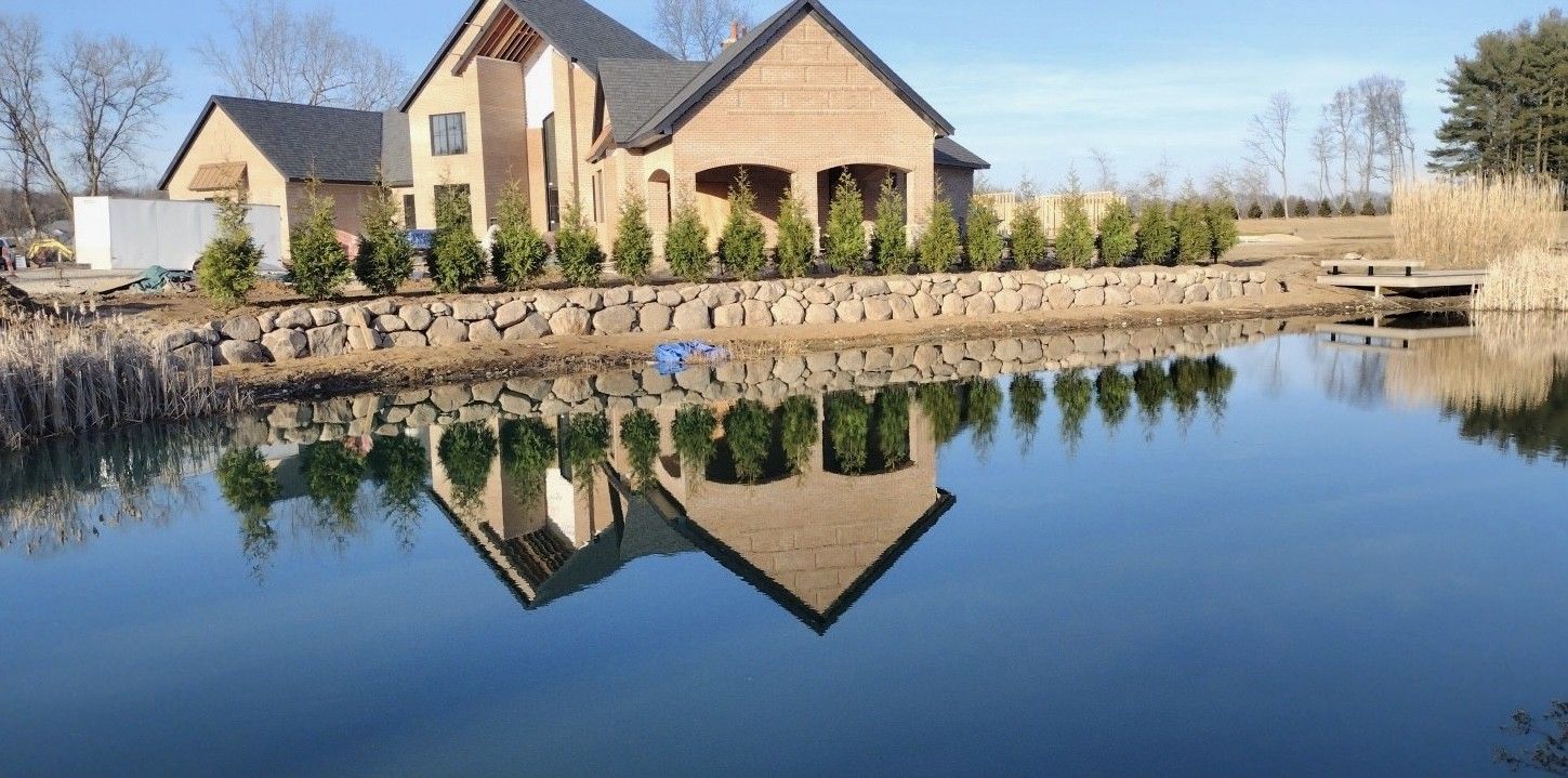 A modern brick house with a dark roof stands behind a row of small trees along a calm pond reflecting the structure.