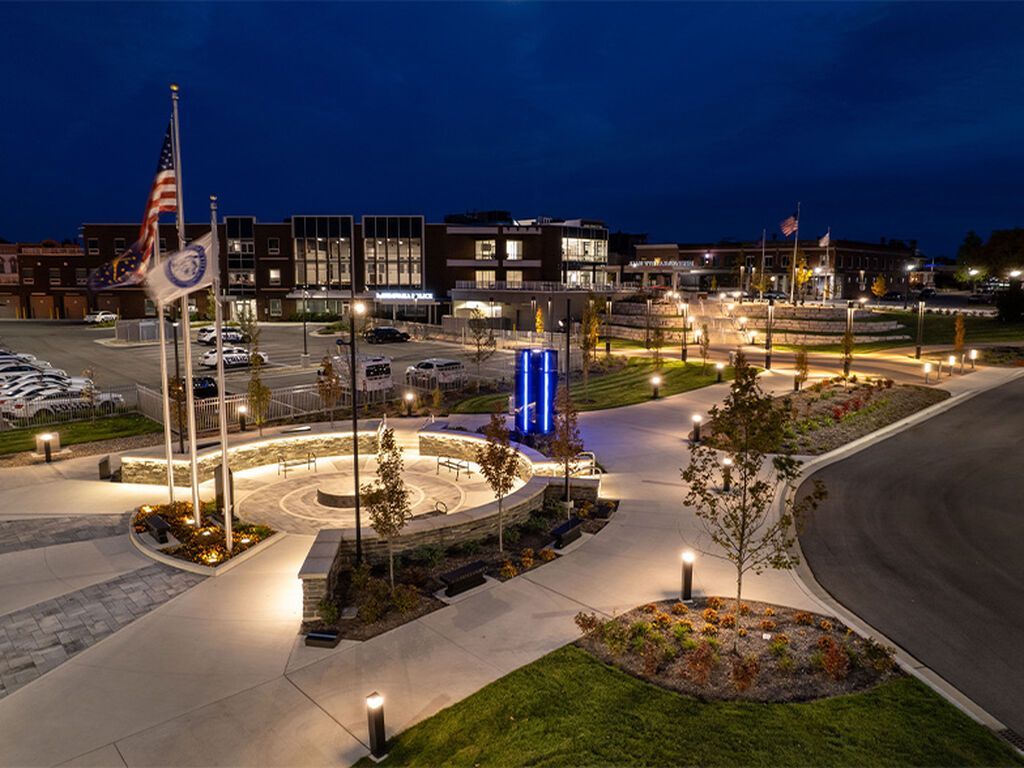 A night scene of a lit memorial plaza with flags, stone walls, and landscaping in front of a modern building.