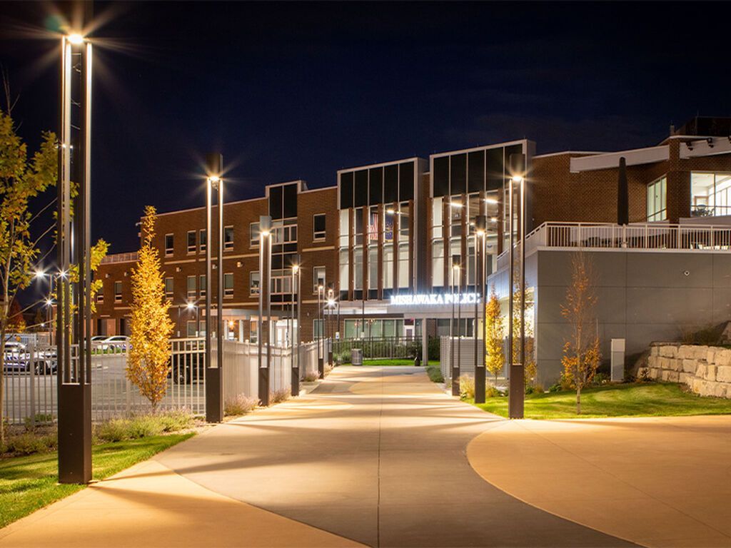 A paved walkway at night lit by modern vertical lamps, leading to the entrance of a contemporary multi-story building.