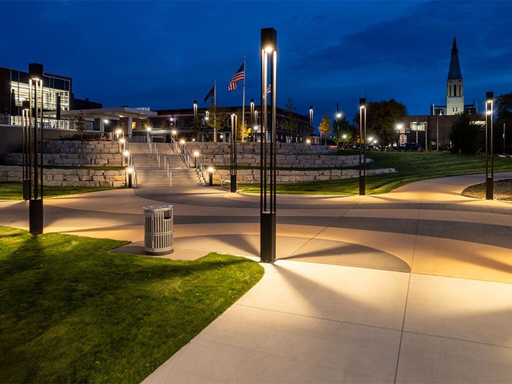 An illuminated outdoor plaza at twilight with modern pathway lighting, stone tiers, and a church steeple in the distance.