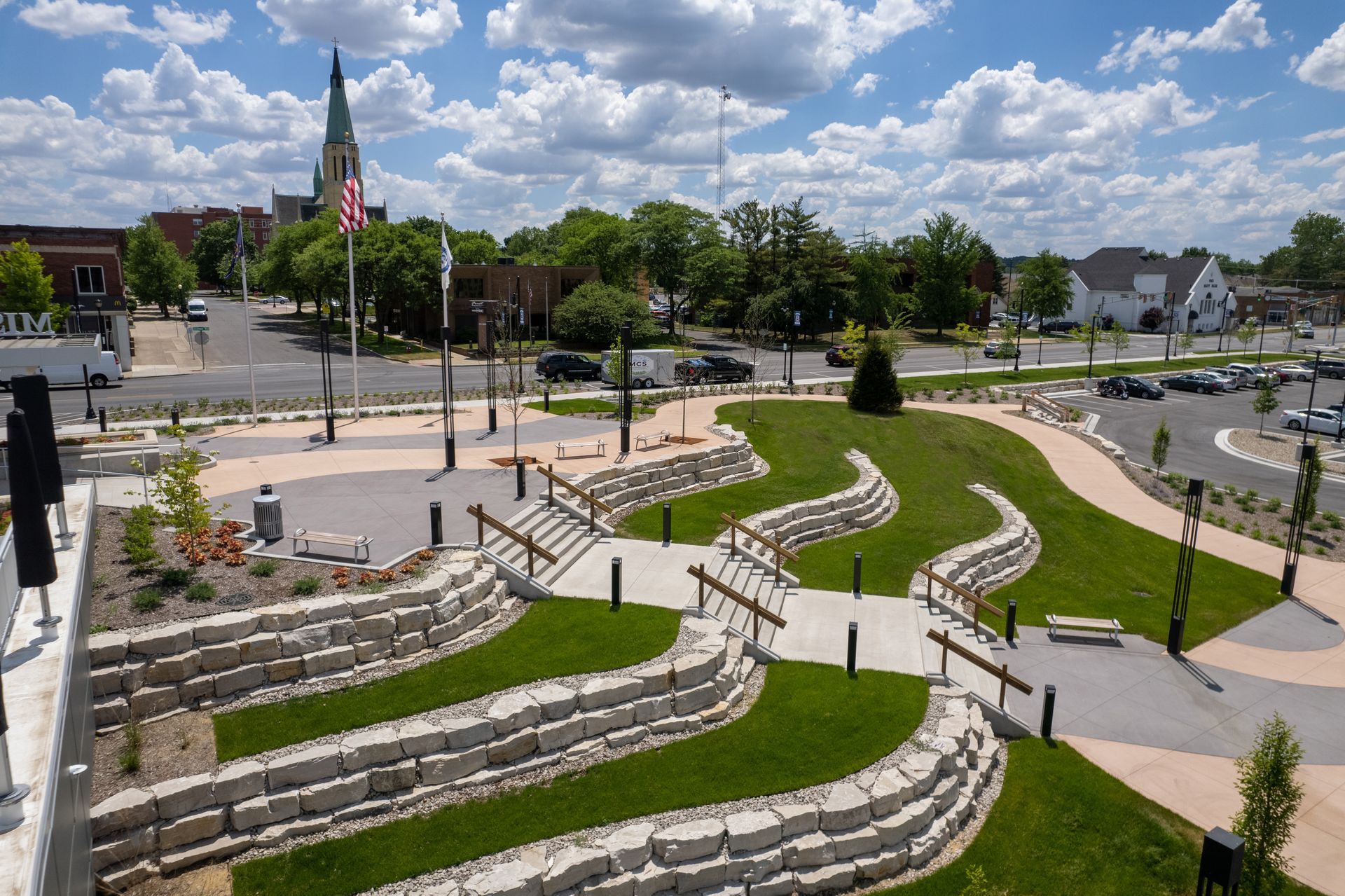 An elevated view of a landscaped public park with terraced stone walls, grass, walkways, and a town church in the background.