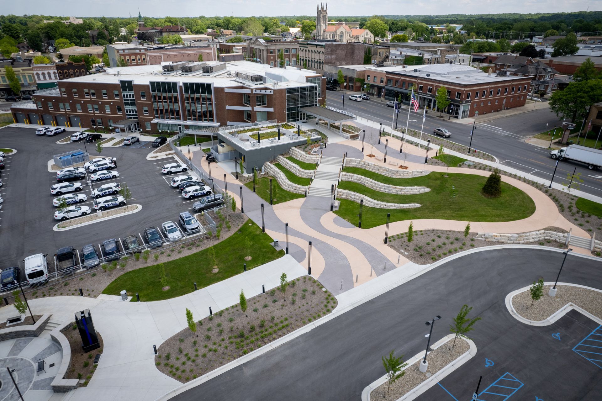 Aerial view of an urban plaza with modern landscaping, paved walkways, and adjacent parking lots near city buildings.