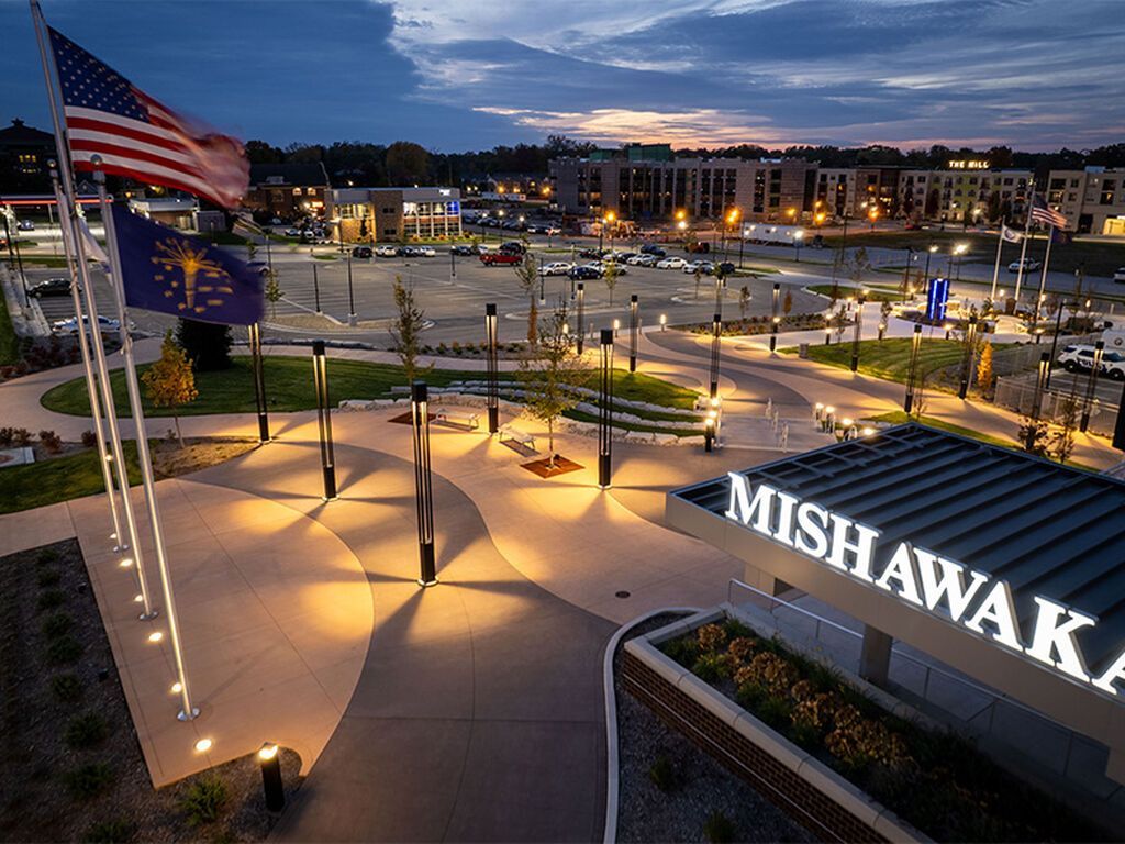 A twilight view of a paved plaza with lighting, landscaping, flags, and a sign reading 