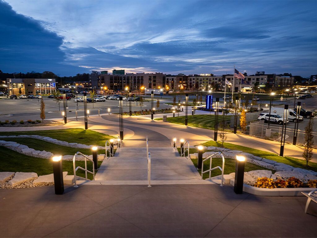 A paved path with railings leads through a landscaped park with glowing light posts toward a commercial area at dusk.