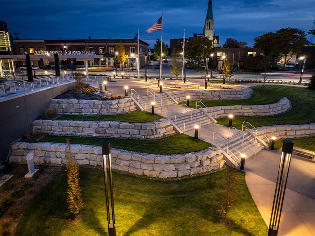A landscaped town square at dusk with tiered stone retaining walls, lit pathways, green grass, and a distant church spire.