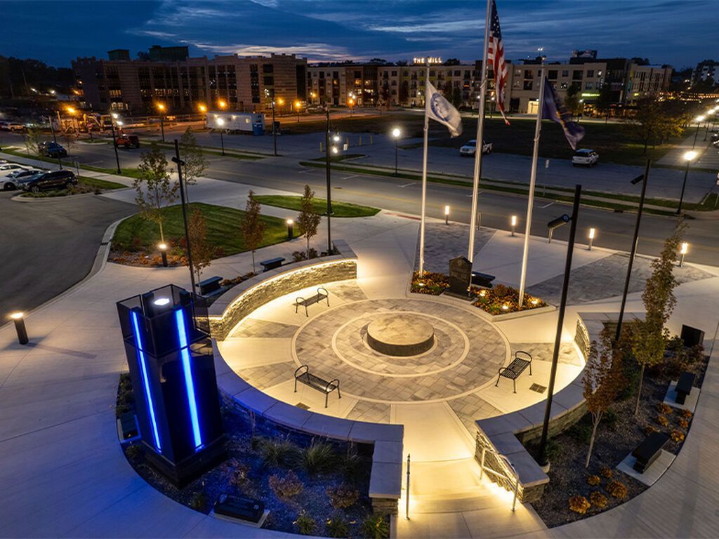 An aerial night view of a lit circular outdoor memorial plaza featuring stone walls, benches, and three flagpoles.