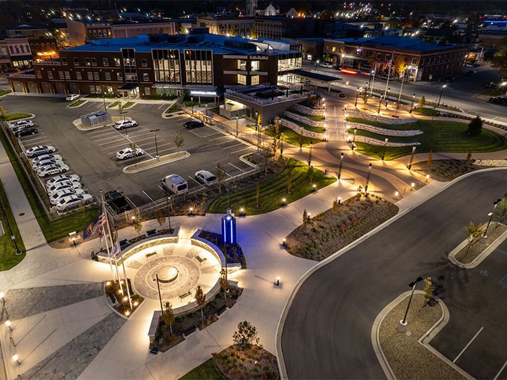 An elevated, night-time view of a illuminated town plaza with a circular memorial, surrounding parking, and buildings.