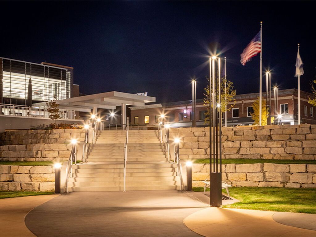 A wide stone staircase at night, illuminated by modern lampposts, leading to an open plaza with American and state flags.