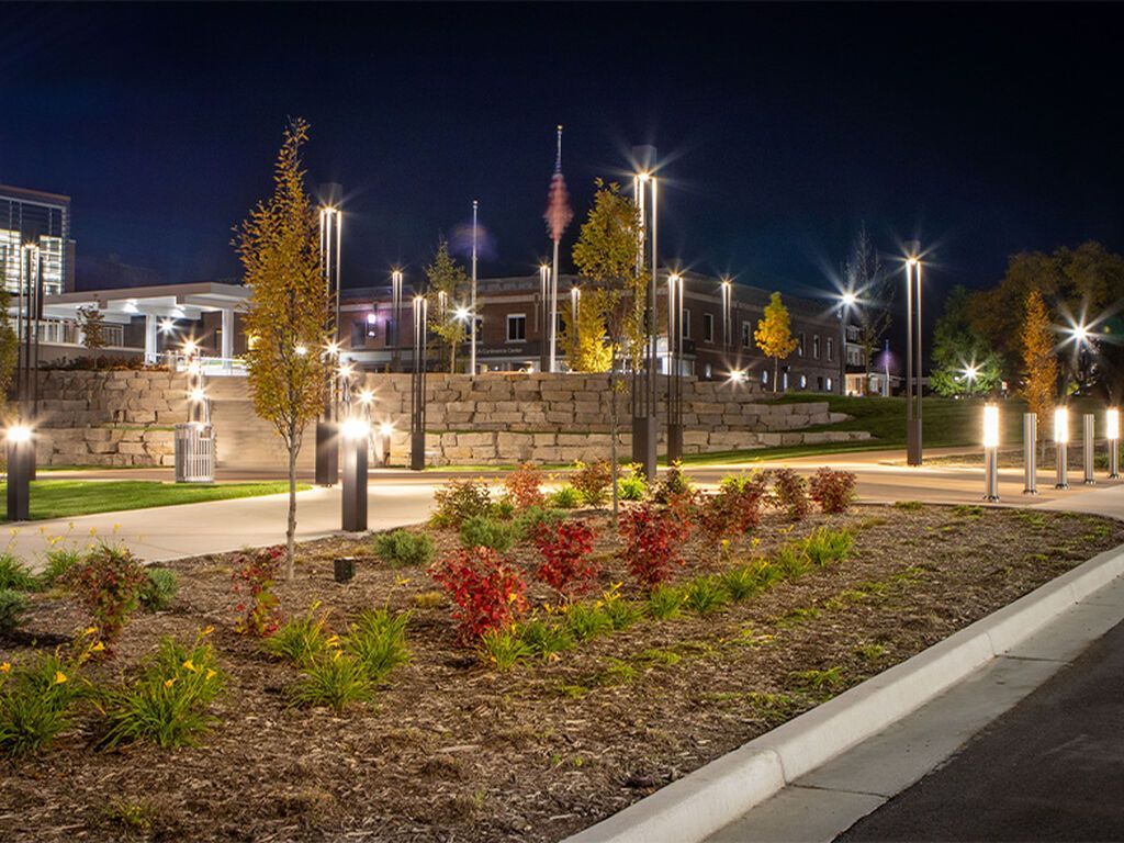 A landscape at night with illuminated trees, garden beds, stone retaining walls, and a paved path lit by street lamps.