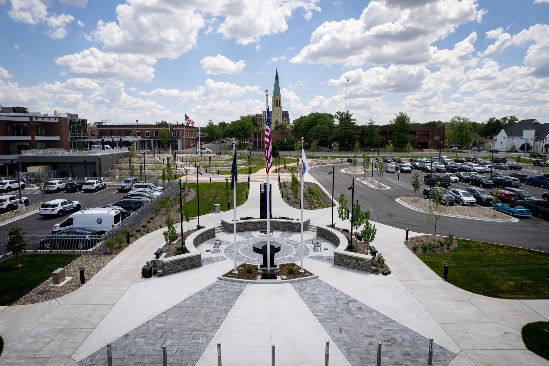 A memorial plaza with a central flag pole and seating area, surrounded by parking lots and a distant church tower.