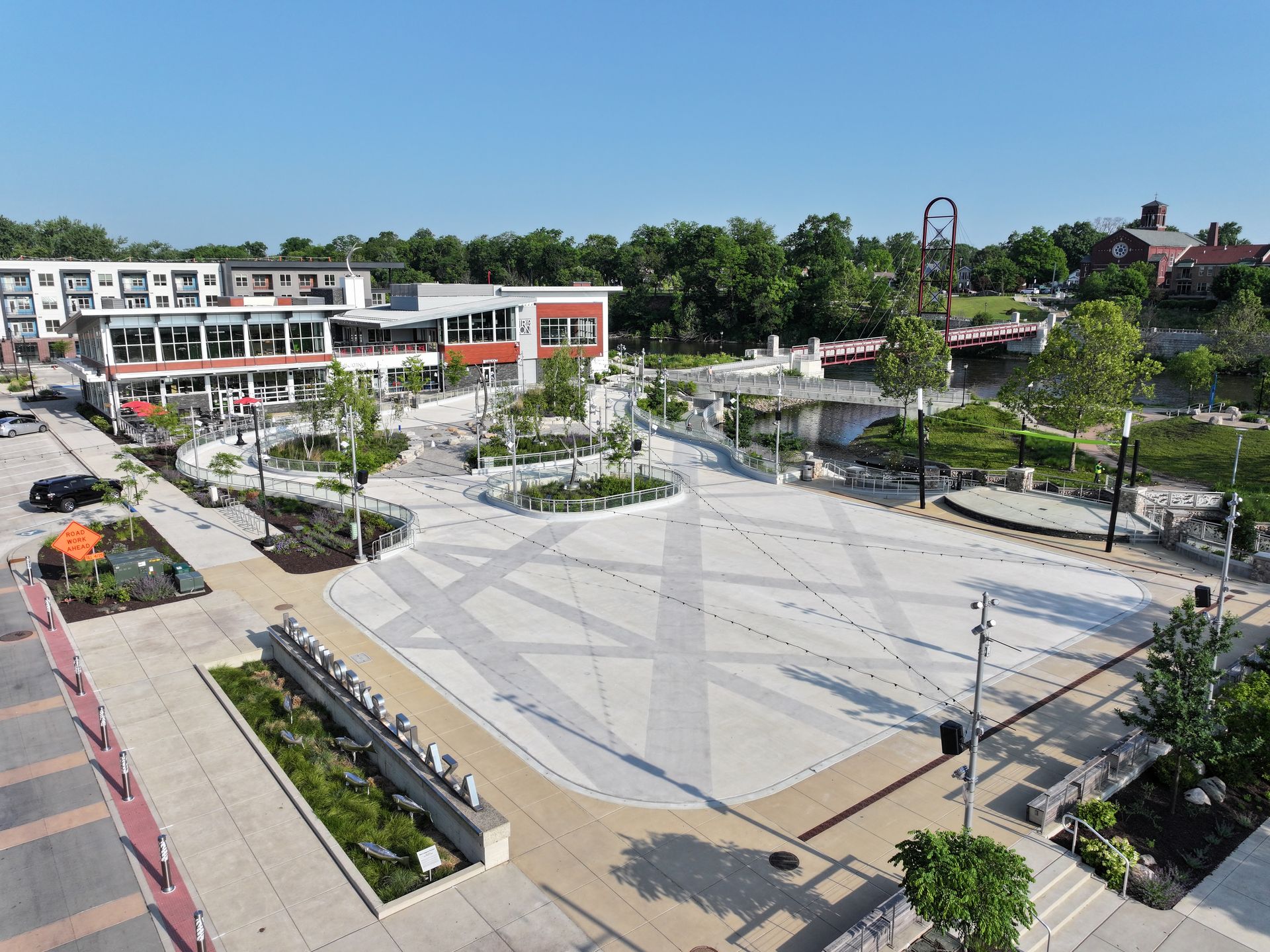 High-angle view of a sunny public plaza with a geometric light-colored tiled floor, surrounding landscaping, and buildings.