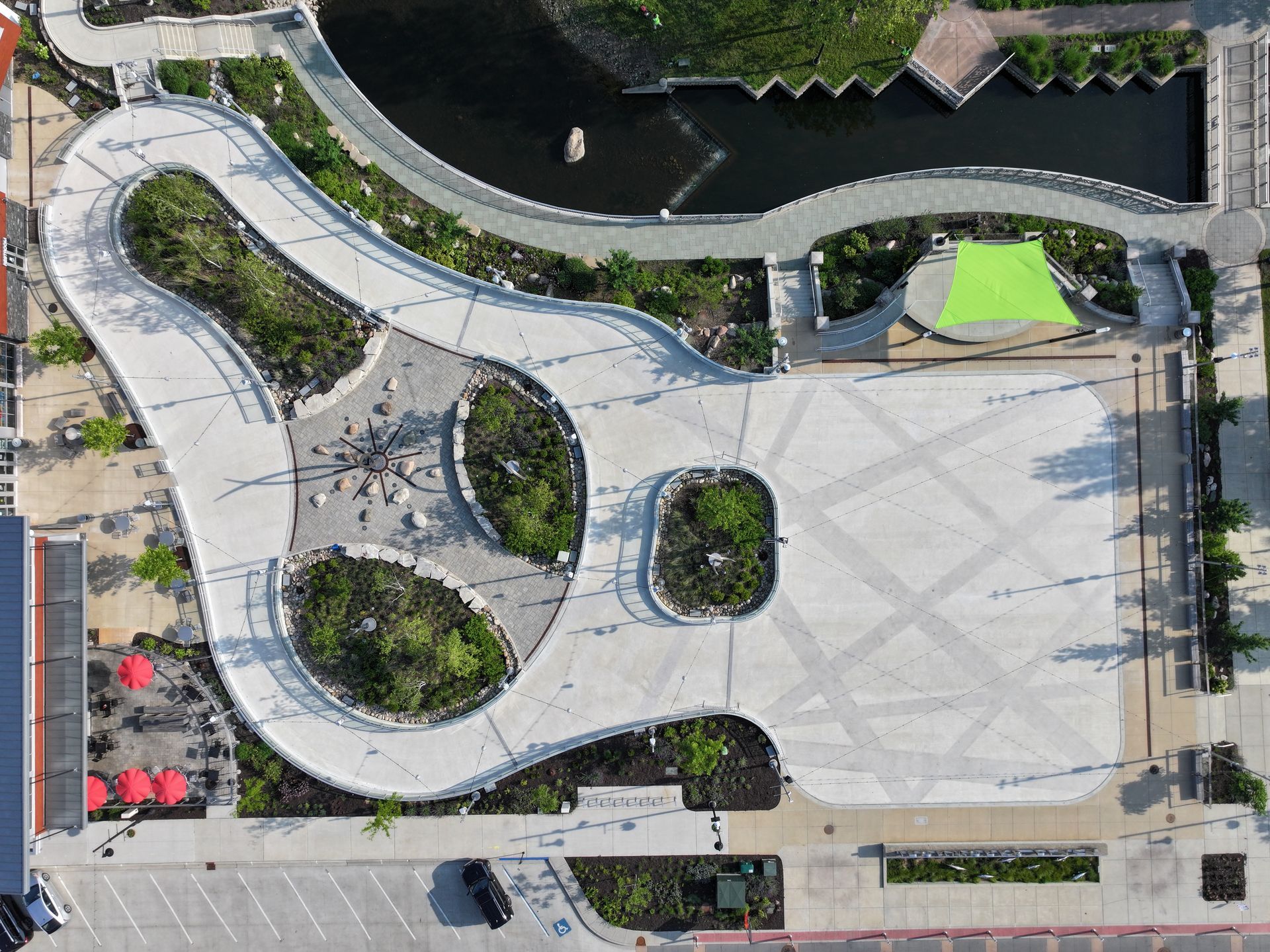 An aerial view of a guitar-shaped concrete plaza with landscaped garden beds, walking paths, and an outdoor stage.