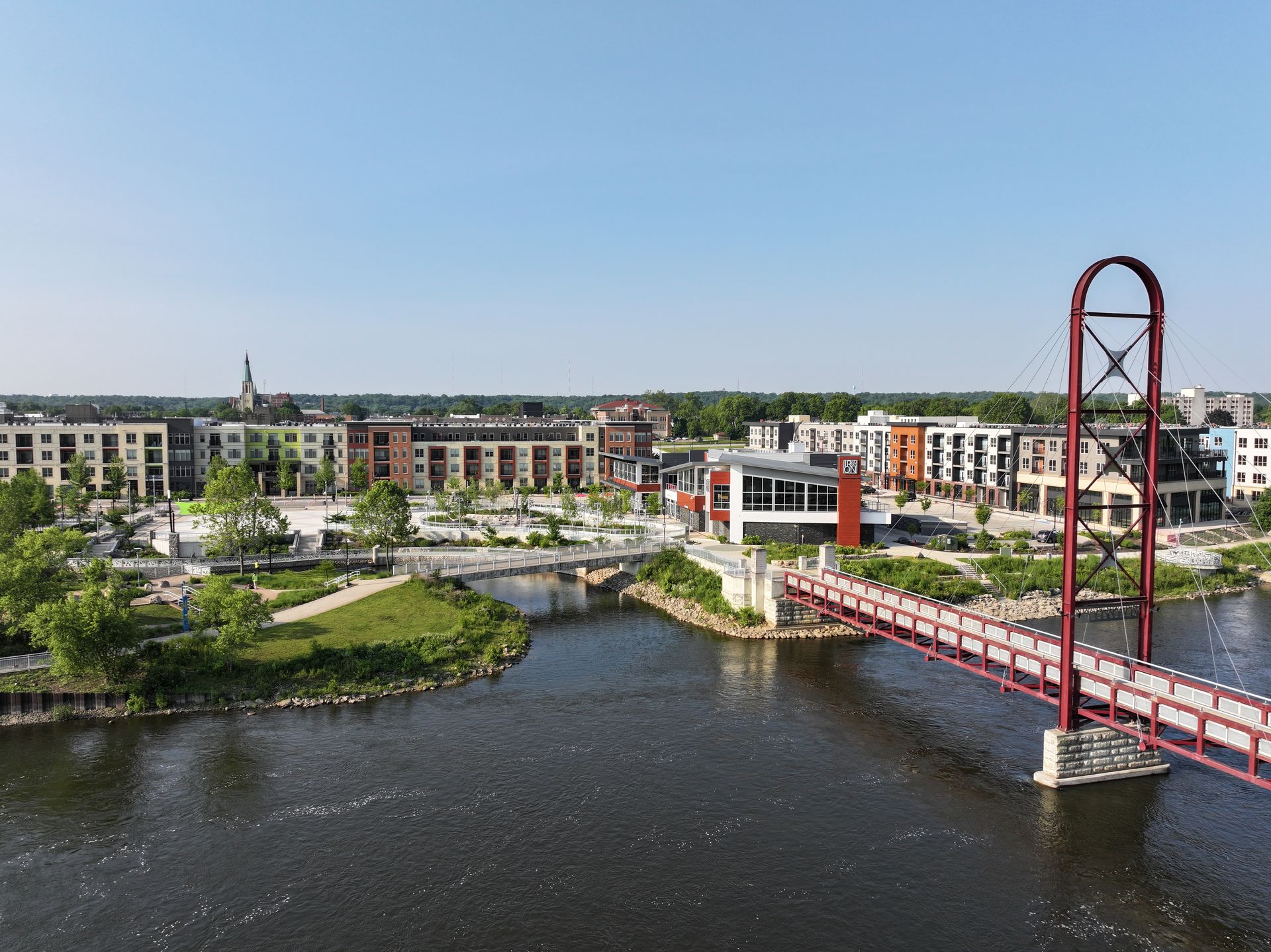 A red cable-stayed pedestrian bridge spans a river leading to a modern apartment complex with trees and walkways.