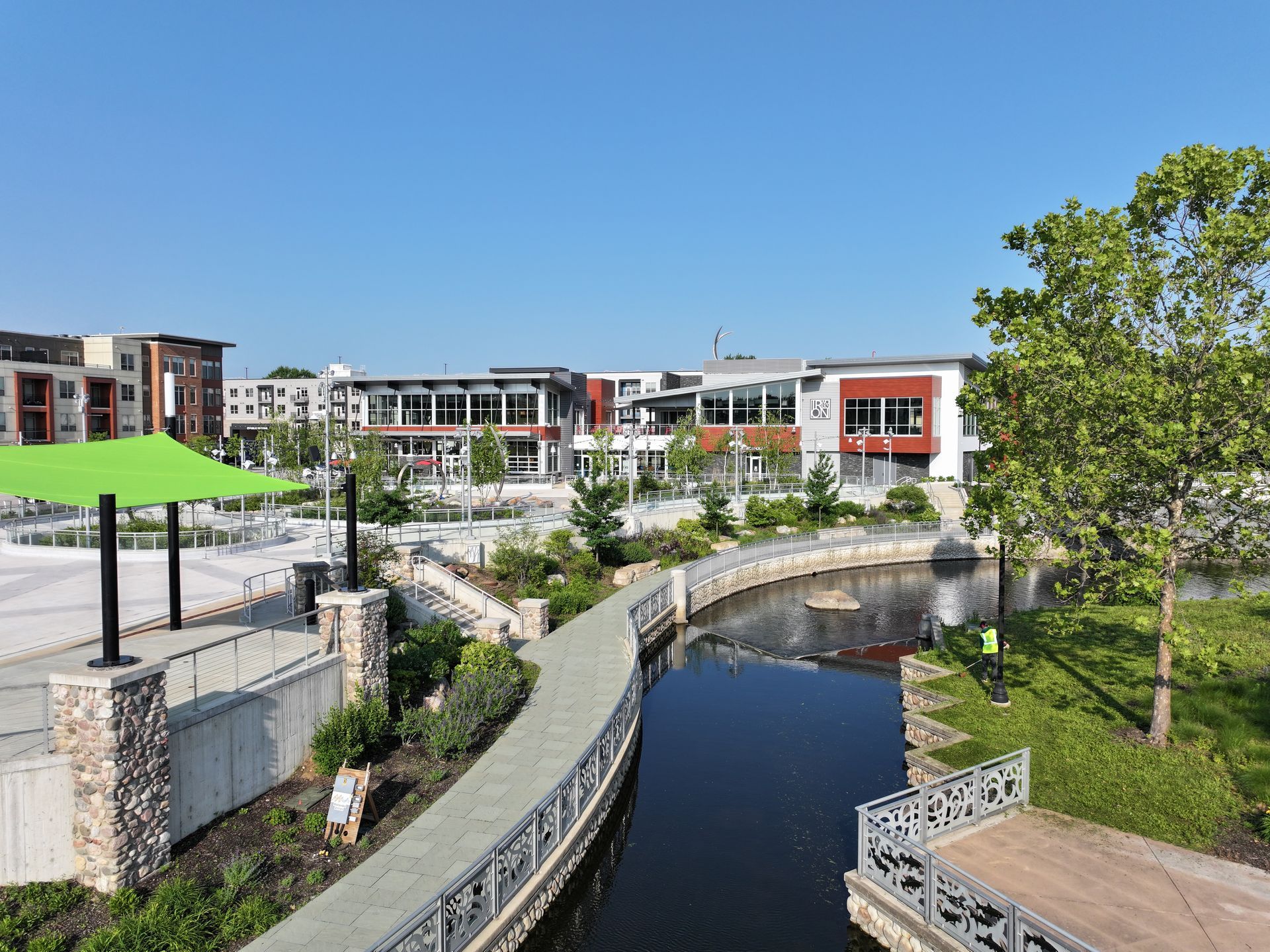 A modern outdoor plaza with a central stream, walking paths, stone accents, and commercial buildings under a blue sky.