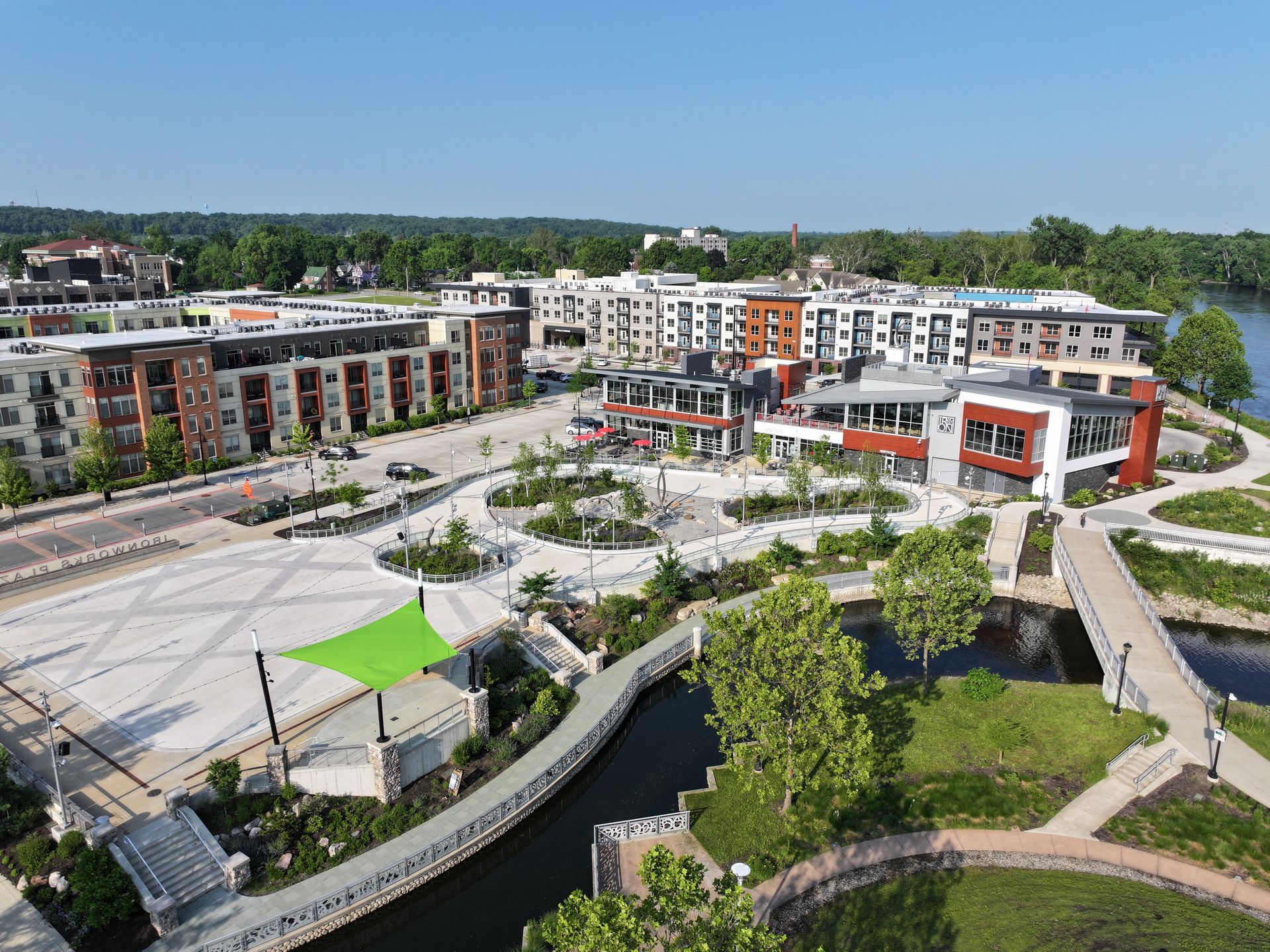 Aerial view of an urban plaza with modern apartment buildings, a green shaded area, and a walkway over a small stream.