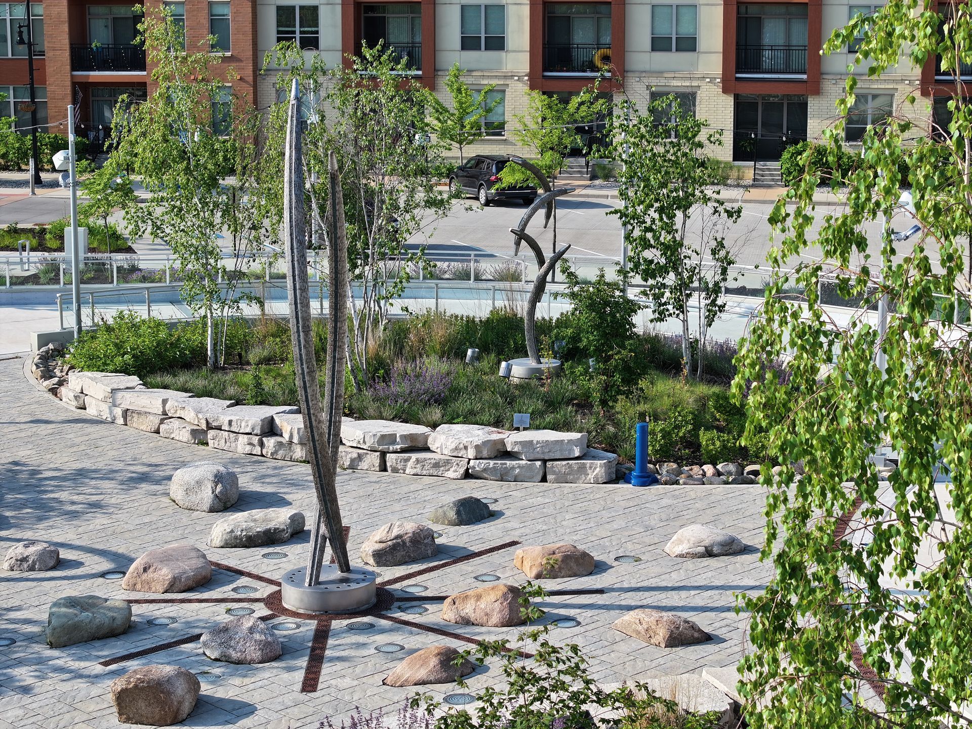A sun-inspired sculpture with radiating metal spikes on a gravel garden path in front of a modern brick apartment complex.