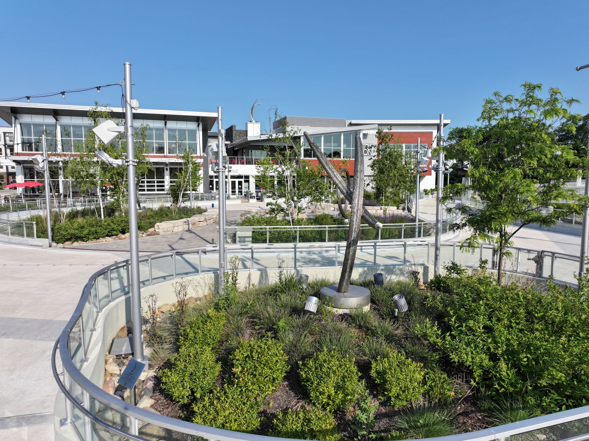 Modern outdoor plaza with a central landscaped planter, a metallic sculpture, and surrounding commercial buildings.