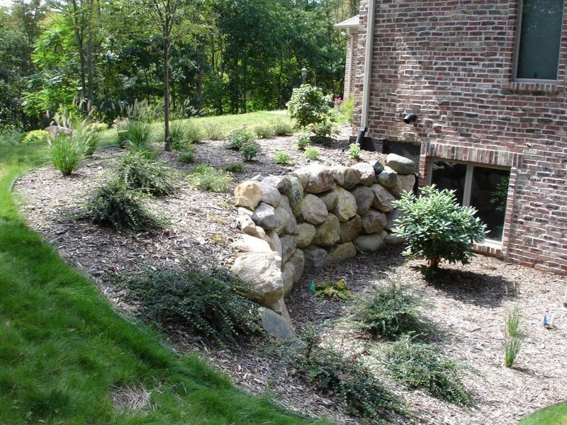 Stone retaining wall on a hillside with plants and mulch, beside a brick building.
