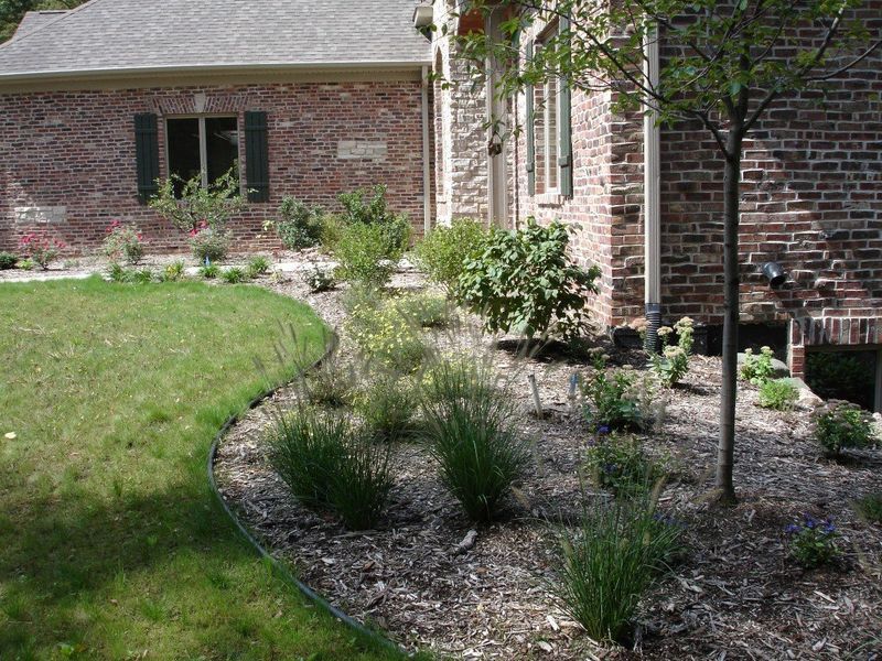 A brick house with a curved garden bed of plants and mulch, bordered by green grass.