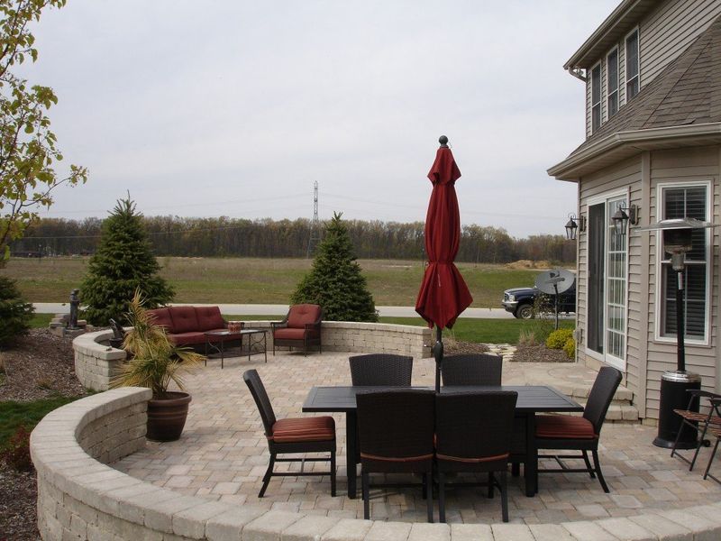 Patio with outdoor furniture, red umbrella, and brick retaining wall, overlooking a field.
