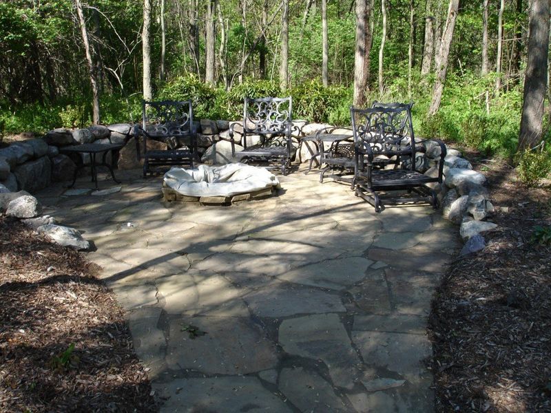 Stone patio with fire pit, surrounded by chairs and trees.