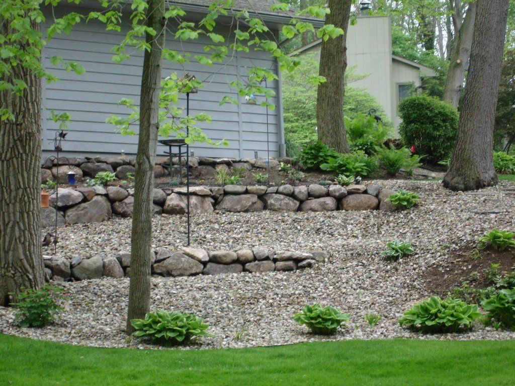 Stone-walled garden beds on a sloped yard, featuring trees and gravel, with a gray garage in the background.