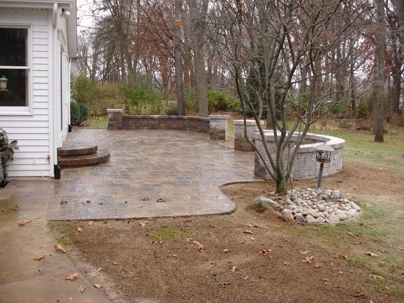Patio with stone pavers, low wall, fire pit, next to a white house, surrounded by trees.
