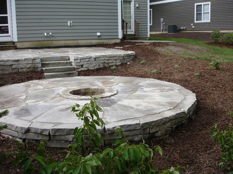 Stone patio with a circular fire pit and retaining walls, in a backyard setting.