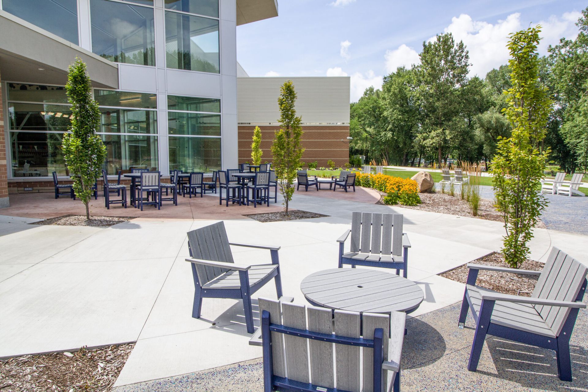 A sunny outdoor patio area featuring gray chairs, round tables, and young trees in front of a modern glass-walled building.