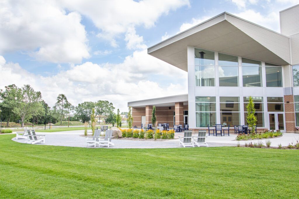 Modern building with large glass windows and a patio featuring lounge chairs, set against a green lawn and cloudy sky.