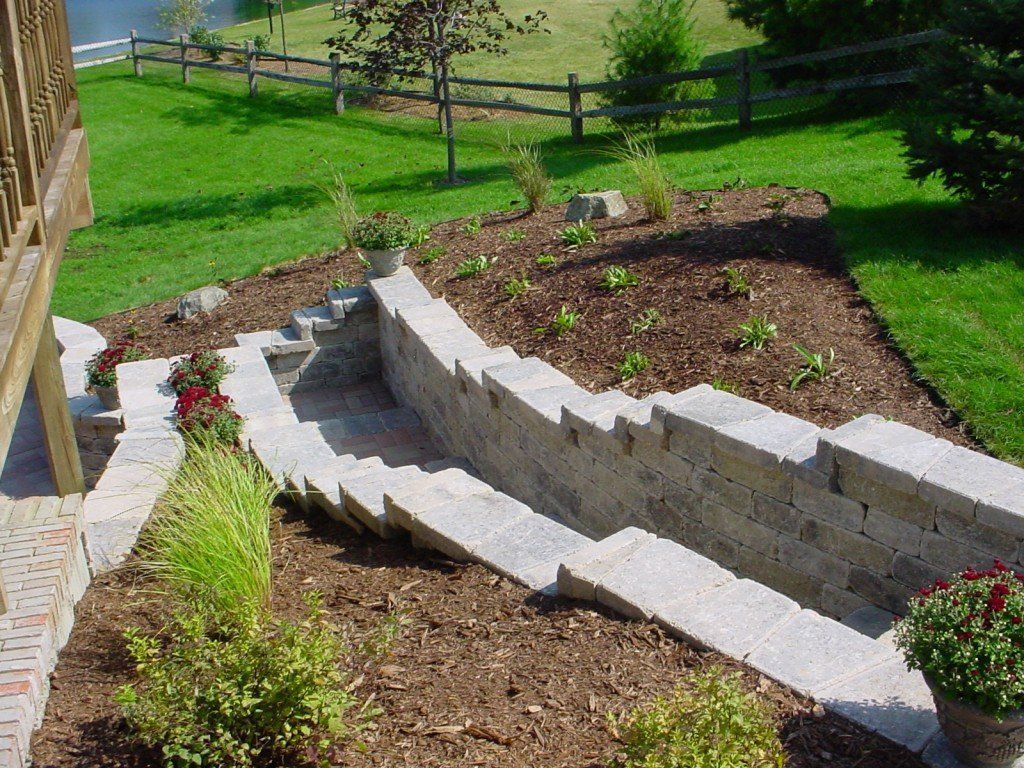Stone steps leading down a sloped yard, bordered by a retaining wall, with flowers and a wooden fence in the background.