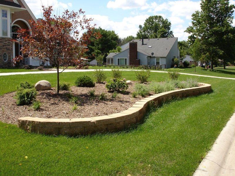 Landscaped yard with a brick-edged flowerbed, trees, and lush green grass on a sunny day.