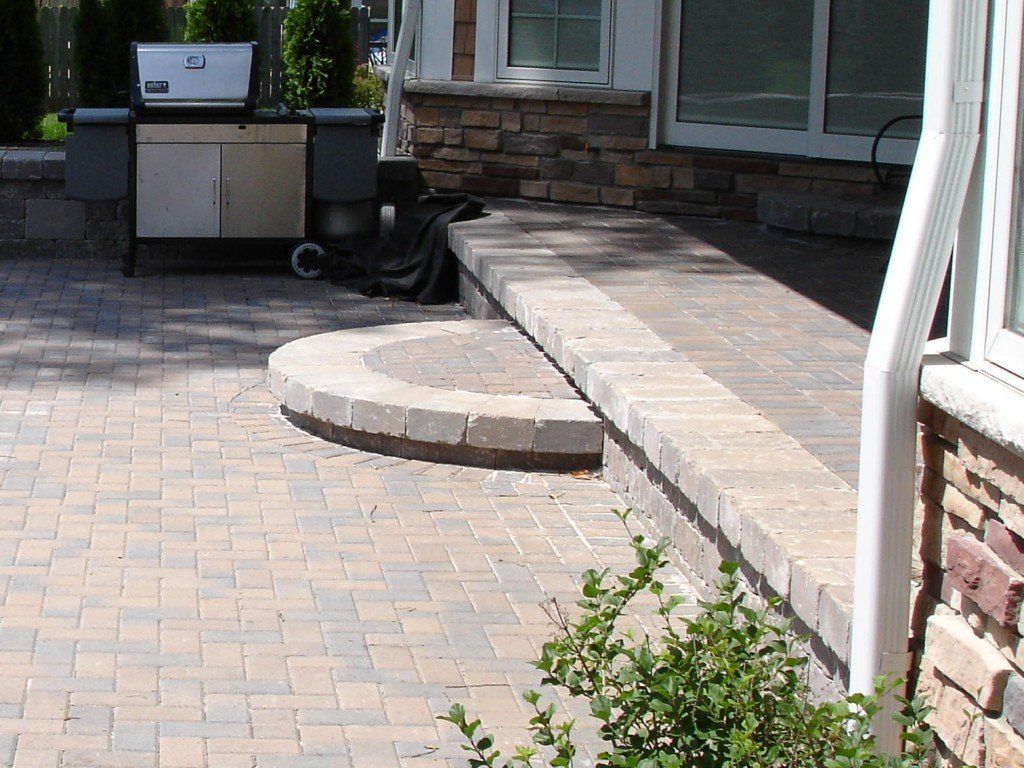Brick patio with a ramp leading up to a house entrance; outdoor grill and shrubbery.