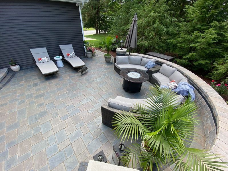 Patio with curved seating, fire pit, and lounge chairs. Dark house exterior and lush greenery surround the outdoor space.