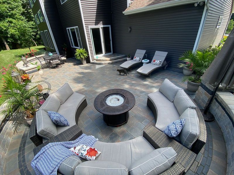 Circular outdoor patio with fire pit, curved sectional sofas, lounge chairs, and house backdrop.