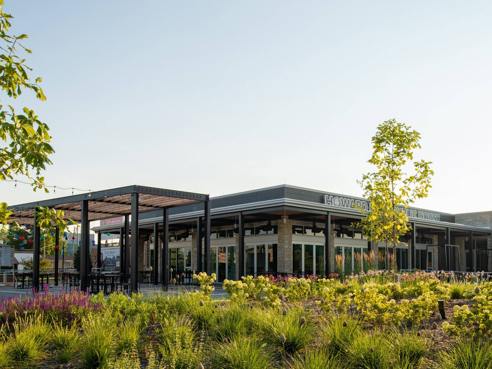 Modern building with a pergola, glass doors, and surrounding landscaping under a bright sky.